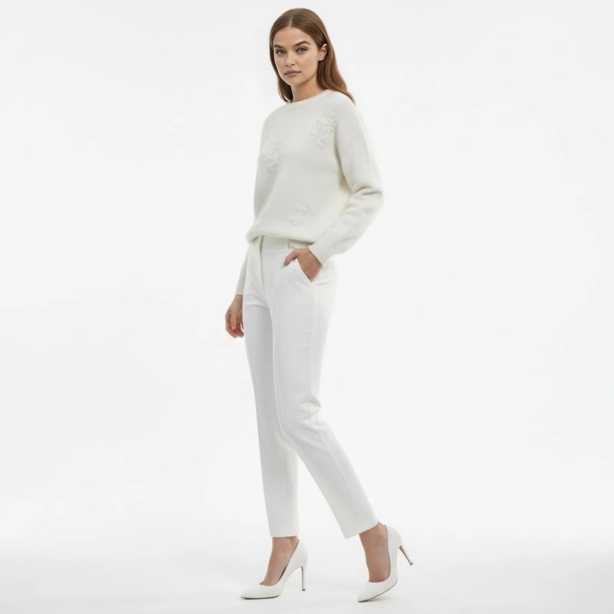 Side profile of a model wearing a white 3D floral embroidered sweater and matching pants with hand in pocket.