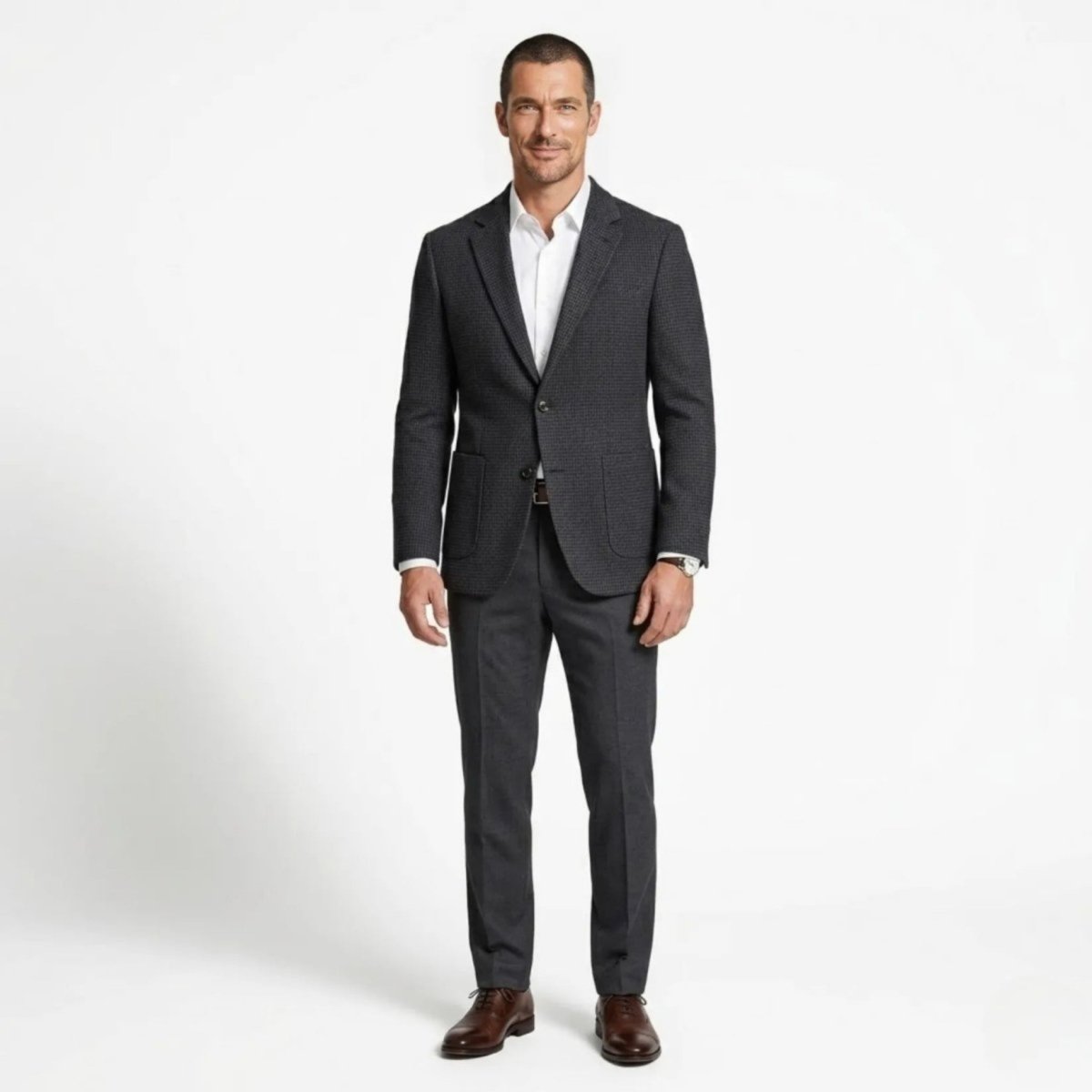 Full body front view of a male model wearing a plaid smart casual suit jacket with grey dress pants and brown shoes.