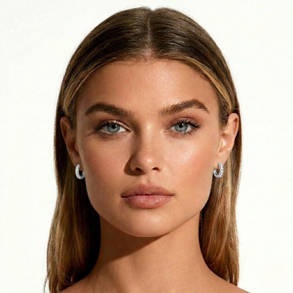 Front-facing portrait of a woman with blonde hair and blue eyes wearing small Kouvr sterling silver diamond huggie earrings, showcasing their size and sparkle.