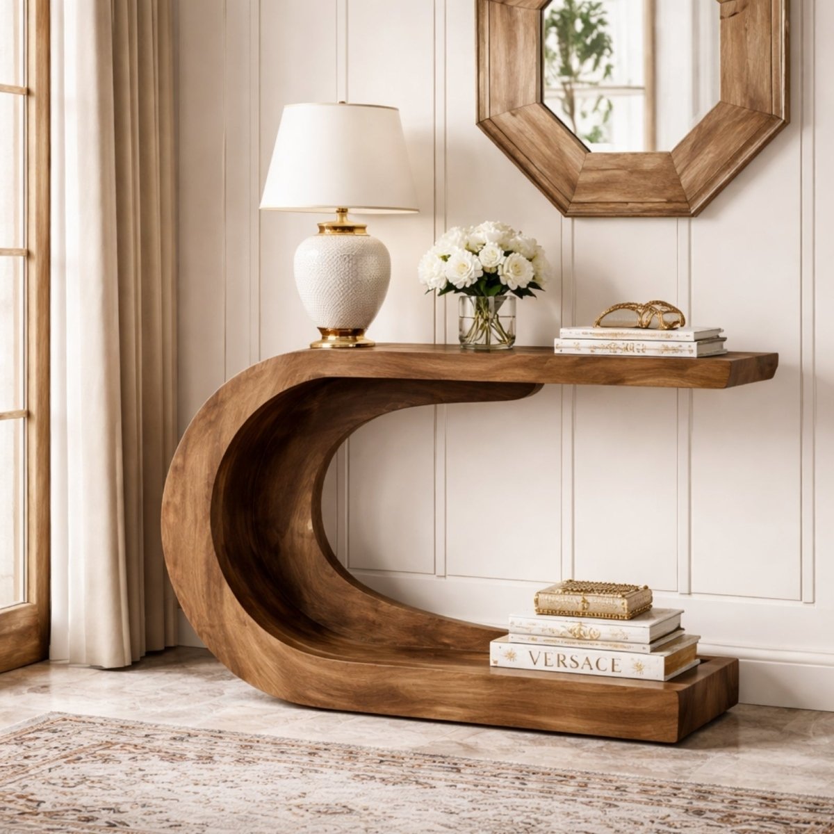 A dark wood C-shaped console table styled with a white lamp, books, and a vase of flowers, placed behind a sofa in a luxury living room setting.