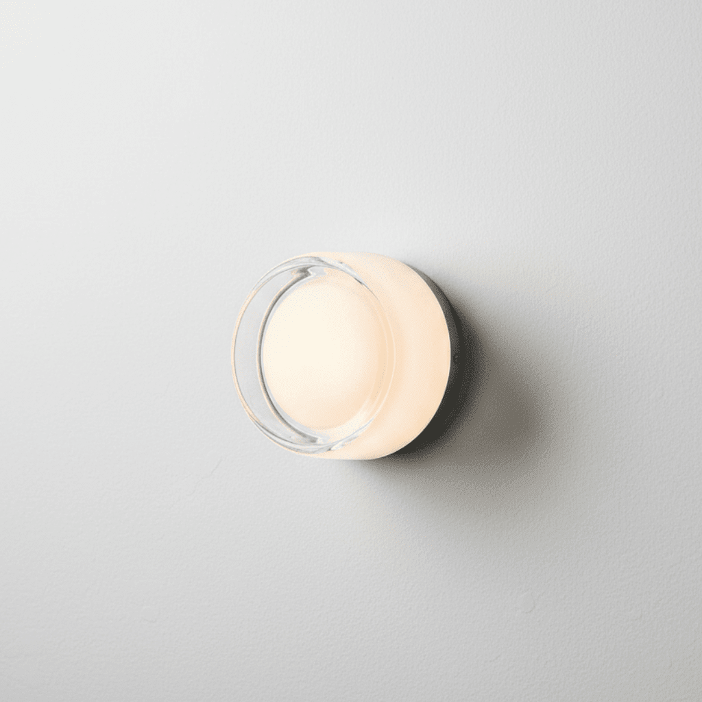 Close-up of a circular minimalist glass wall sconce emitting a warm amber glow against a textured beige plaster wall.