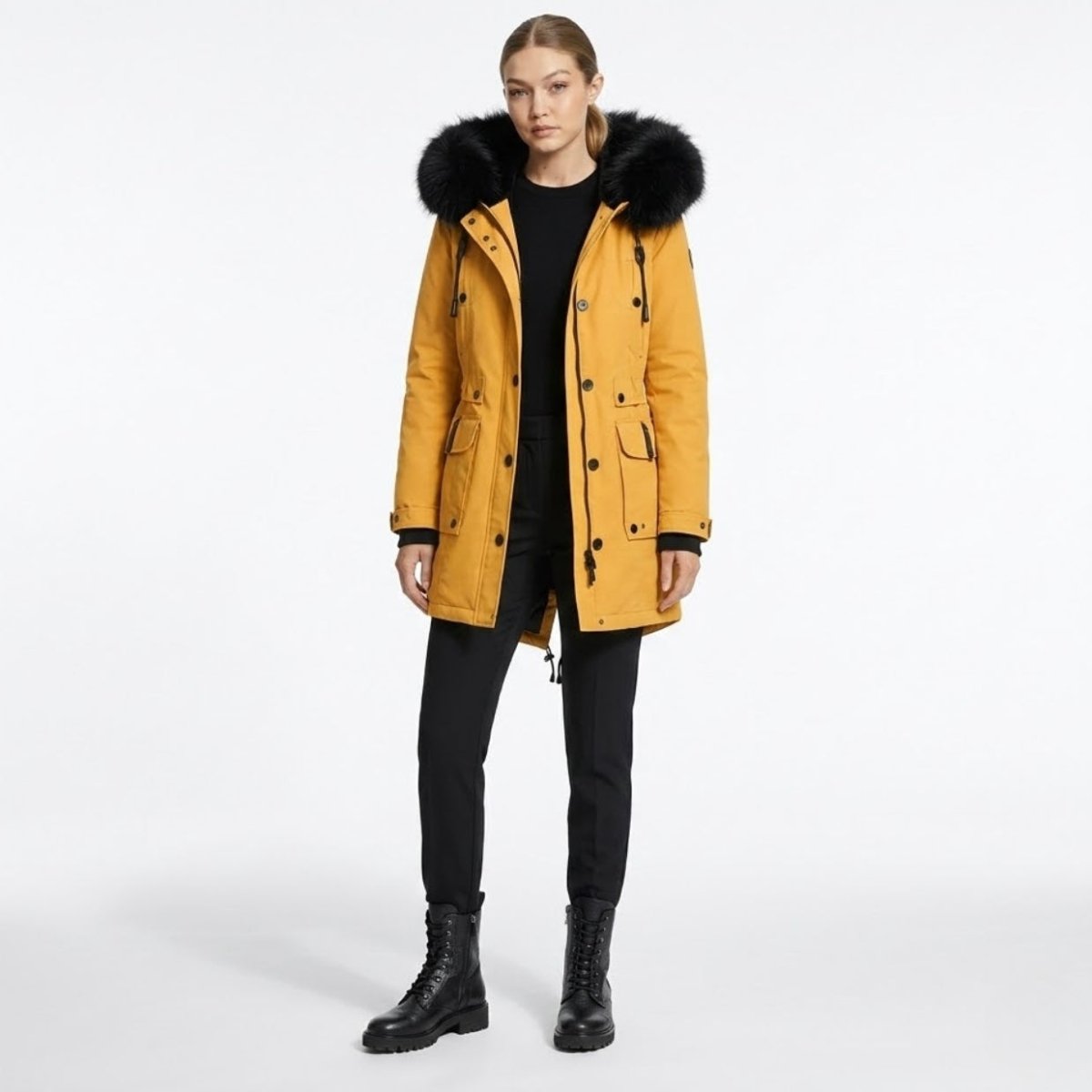 Full length front view of a female model wearing a Kouvr mustard yellow winter parka with a voluminous black faux fur hood, paired with black pants and boots.