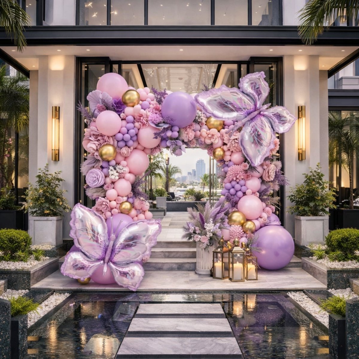 An outdoor garden party balloon arch featuring pampas grass, custom floral arrangements, large matte lavender balloons, and giant iridescent butterfly balloons for a whimsical nature theme