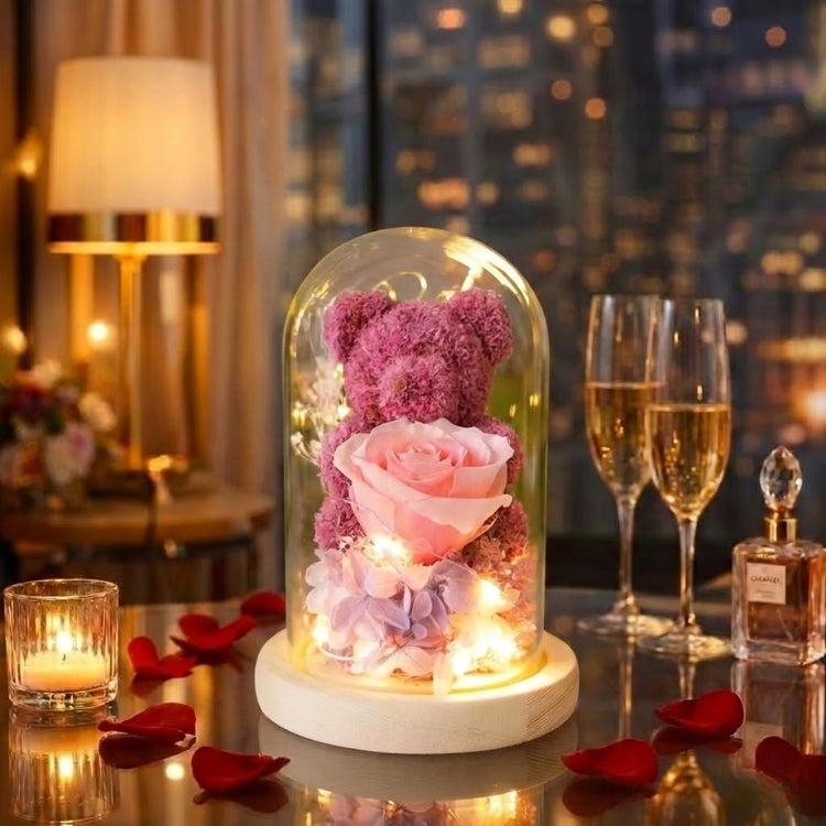 Pink preserved moss teddy bear holding a pink rose in a light-up glass dome against a blurred city night skyline.