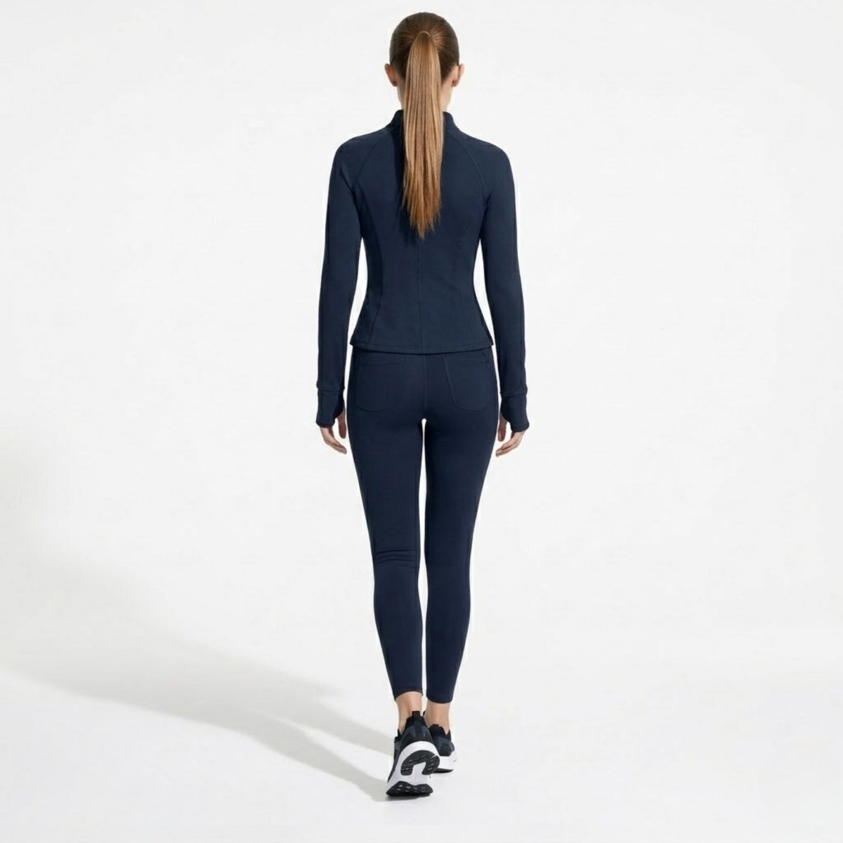 Full body back view of woman walking in fitted navy blue athleisure jacket and leggings
