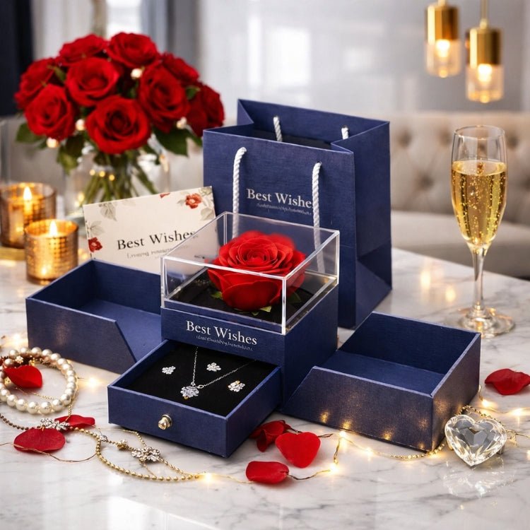 Blue 'Best Wishes' gift box featuring a preserved red rose in a clear case and a pull-out jewelry drawer containing a silver necklace and earrings, set on a marble table with roses and candles.