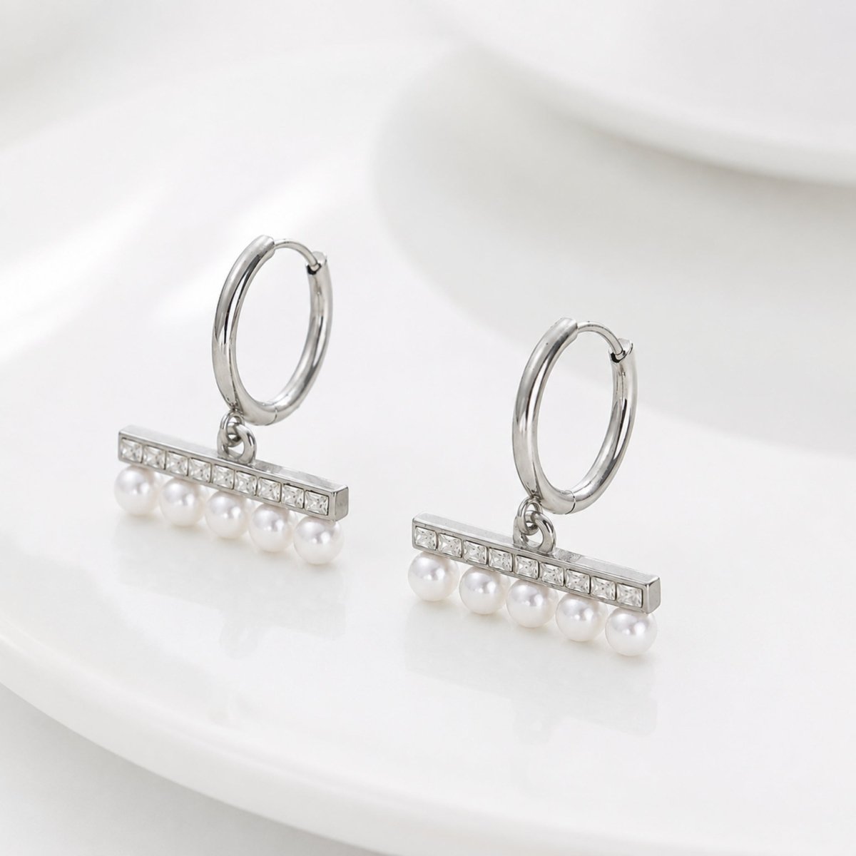 Elegant silver finish huggie hoop earrings featuring a sparkling crystal bar and dainty dangling pearls resting on a clean white surface.