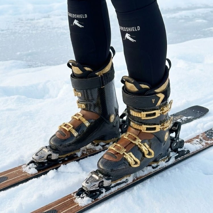 A close-up shot of a skier's legs wearing ski boots and black ShredShield shin protectors, standing on skis in fresh snow.