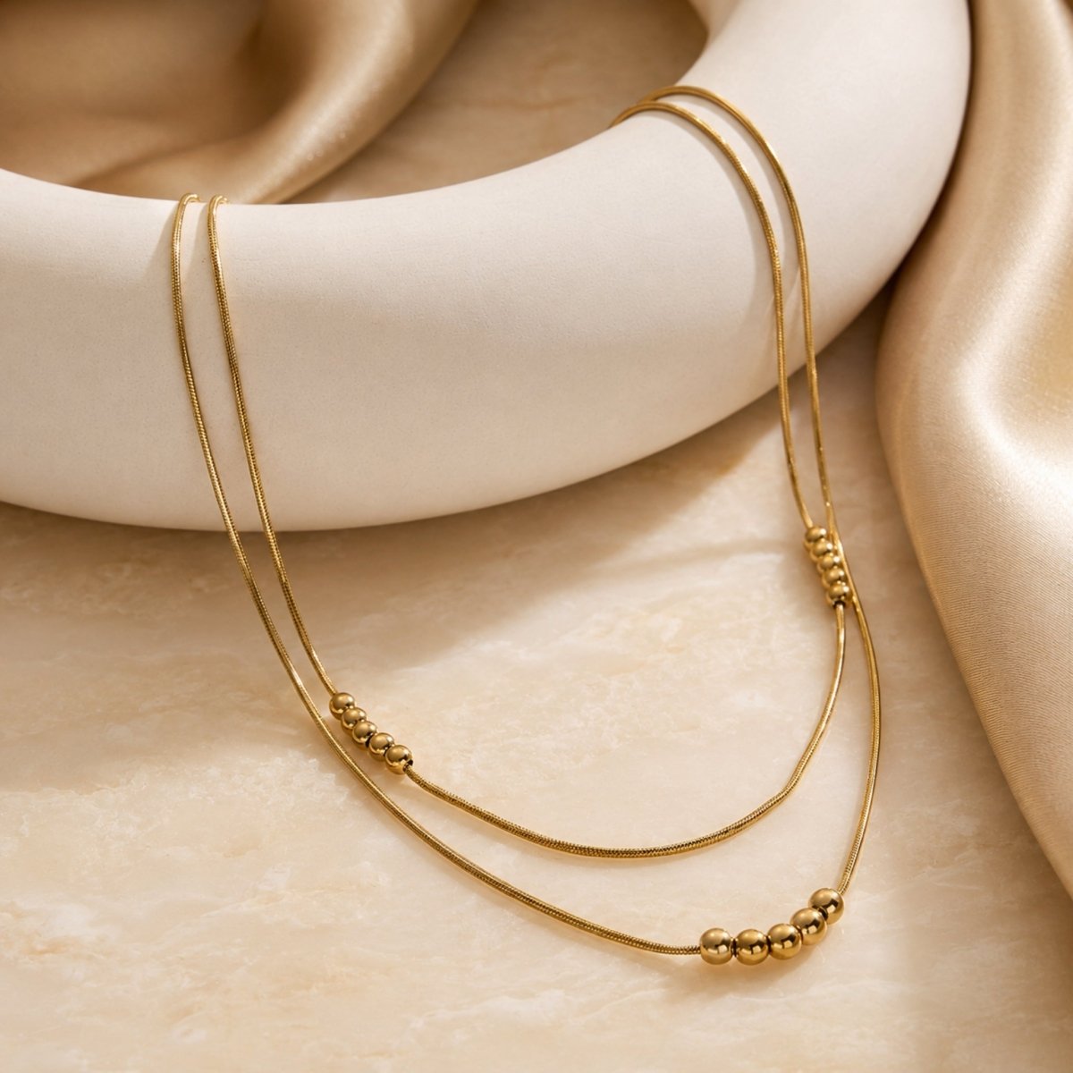 Luxury gold double layered metal bead necklace resting beautifully on a modern white ceramic ring prop and draped beige silk fabric.