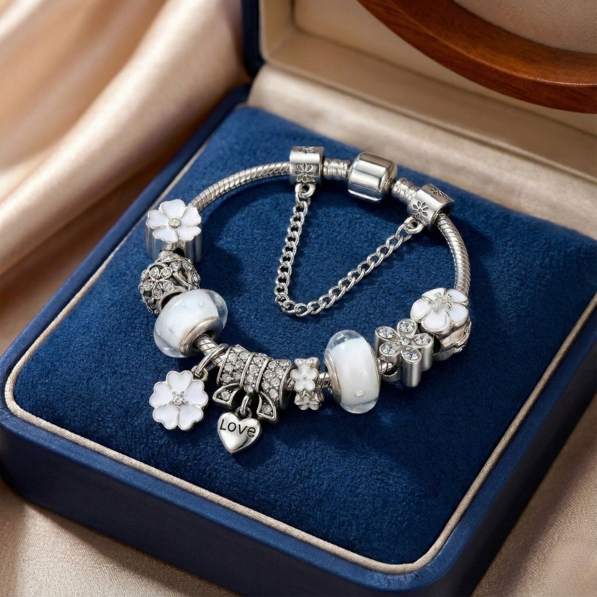 Silver charm bracelet featuring white enamel flower charms, white Murano glass beads, and a safety chain displayed in a blue presentation box.