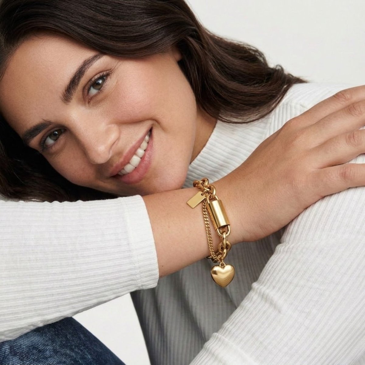 A smiling woman wearing a gold chunky heart charm bracelet, styled with a white ribbed top for a lifestyle fashion look.
