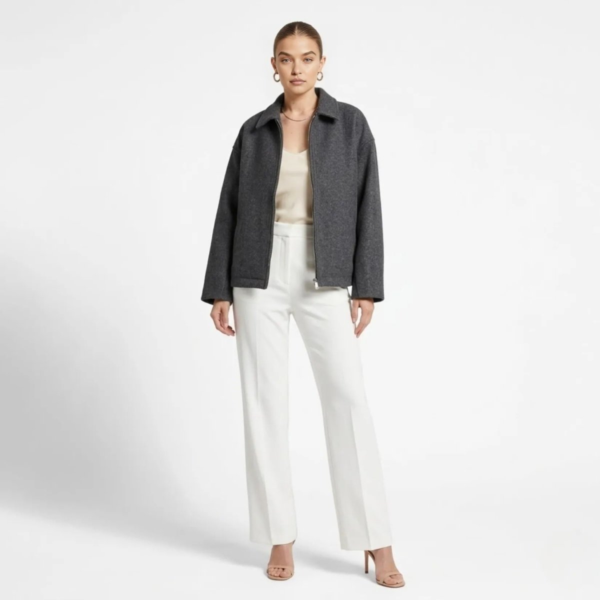 Front view of female model wearing a grey boxy wool blend zip up collared jacket by Kouvr styled with white high waisted trousers.