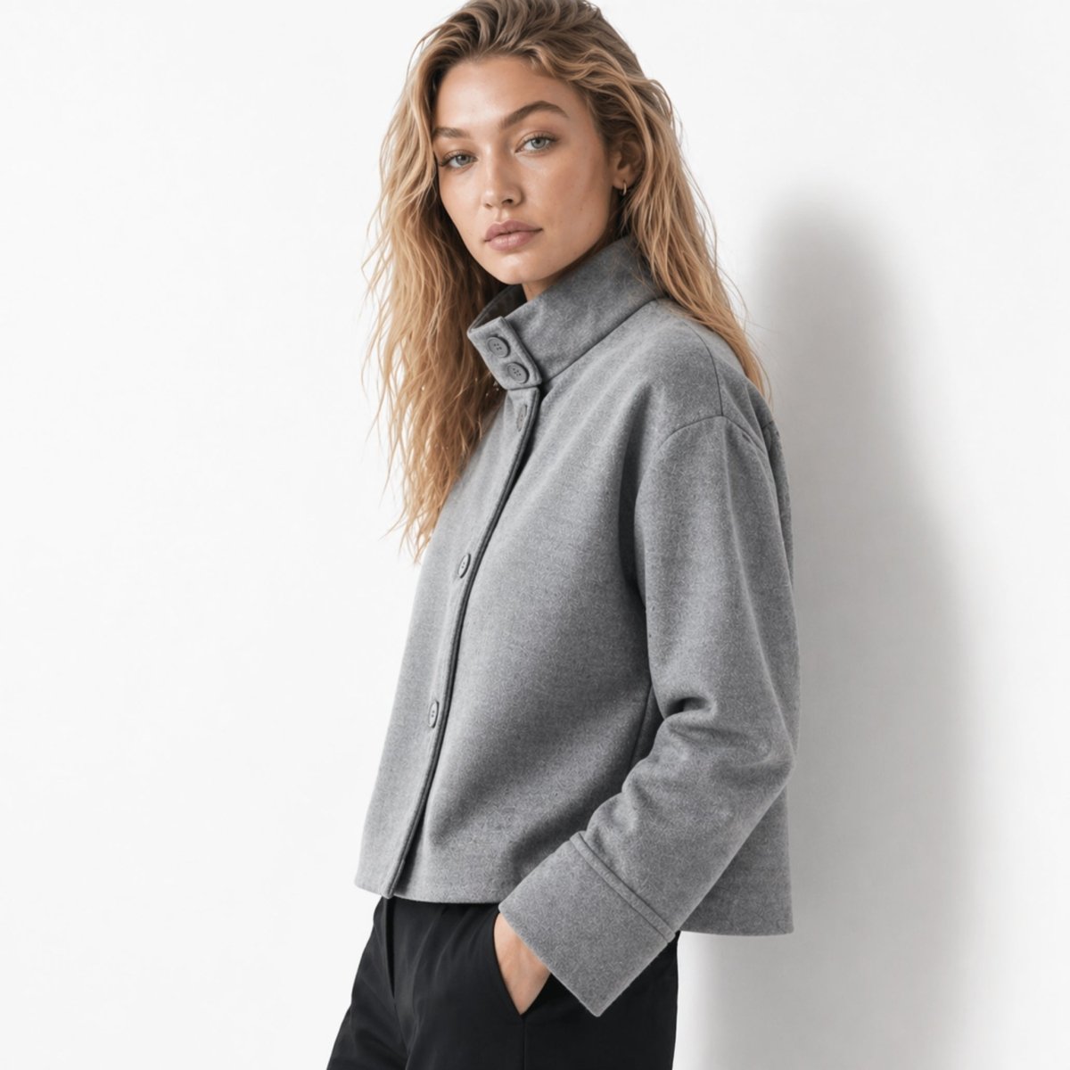 Side angle of a woman modeling a minimalist grey cropped jacket with wide sleeves and a high collar.