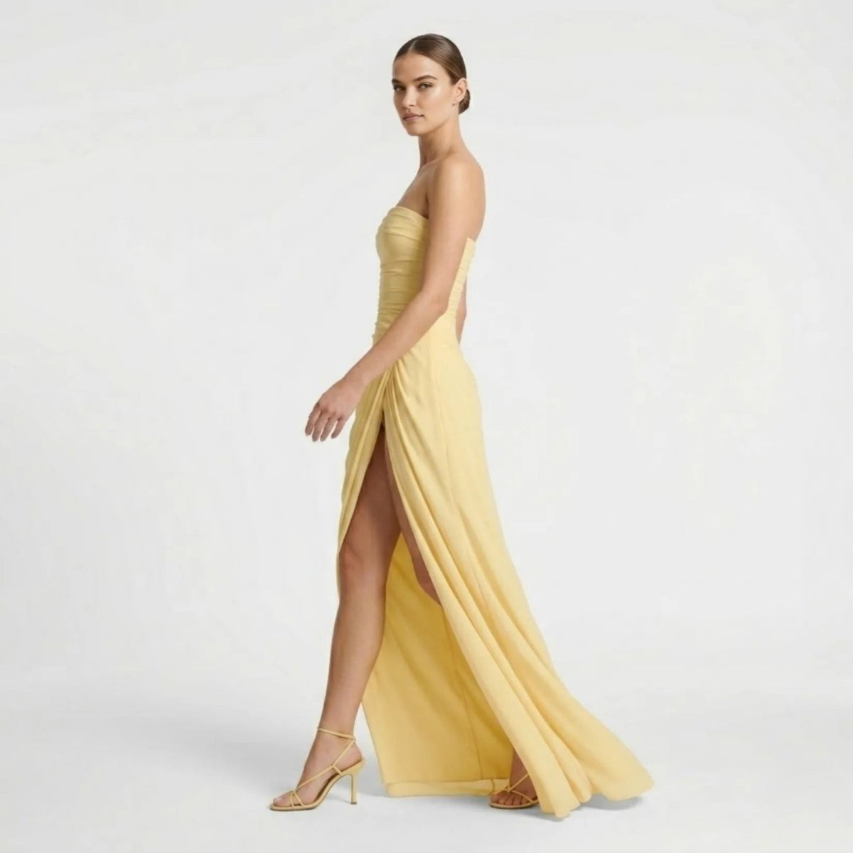 Side profile of fashion model in yellow corset back evening gown showing high leg slit and strappy heels.