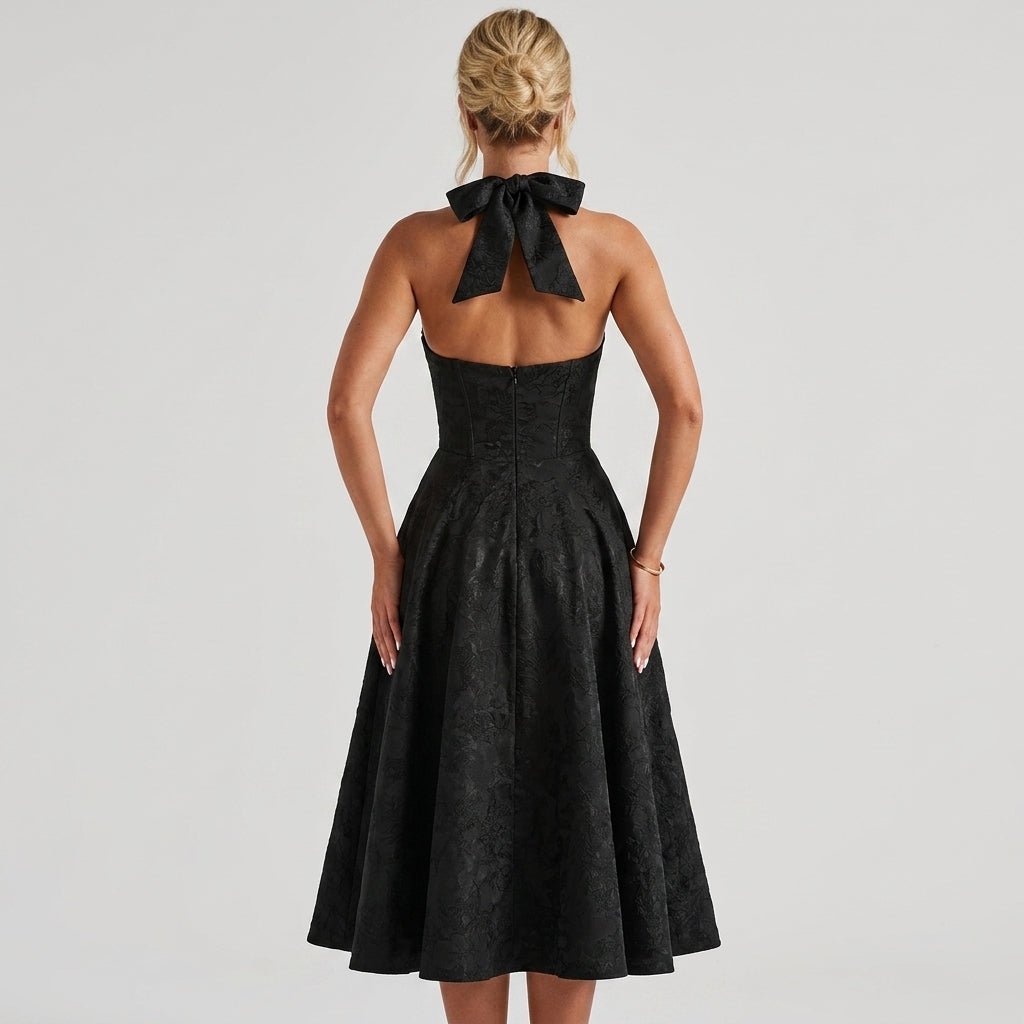 Back view of black fit and flare midi dress showing oversized halter bow tie and open back design.