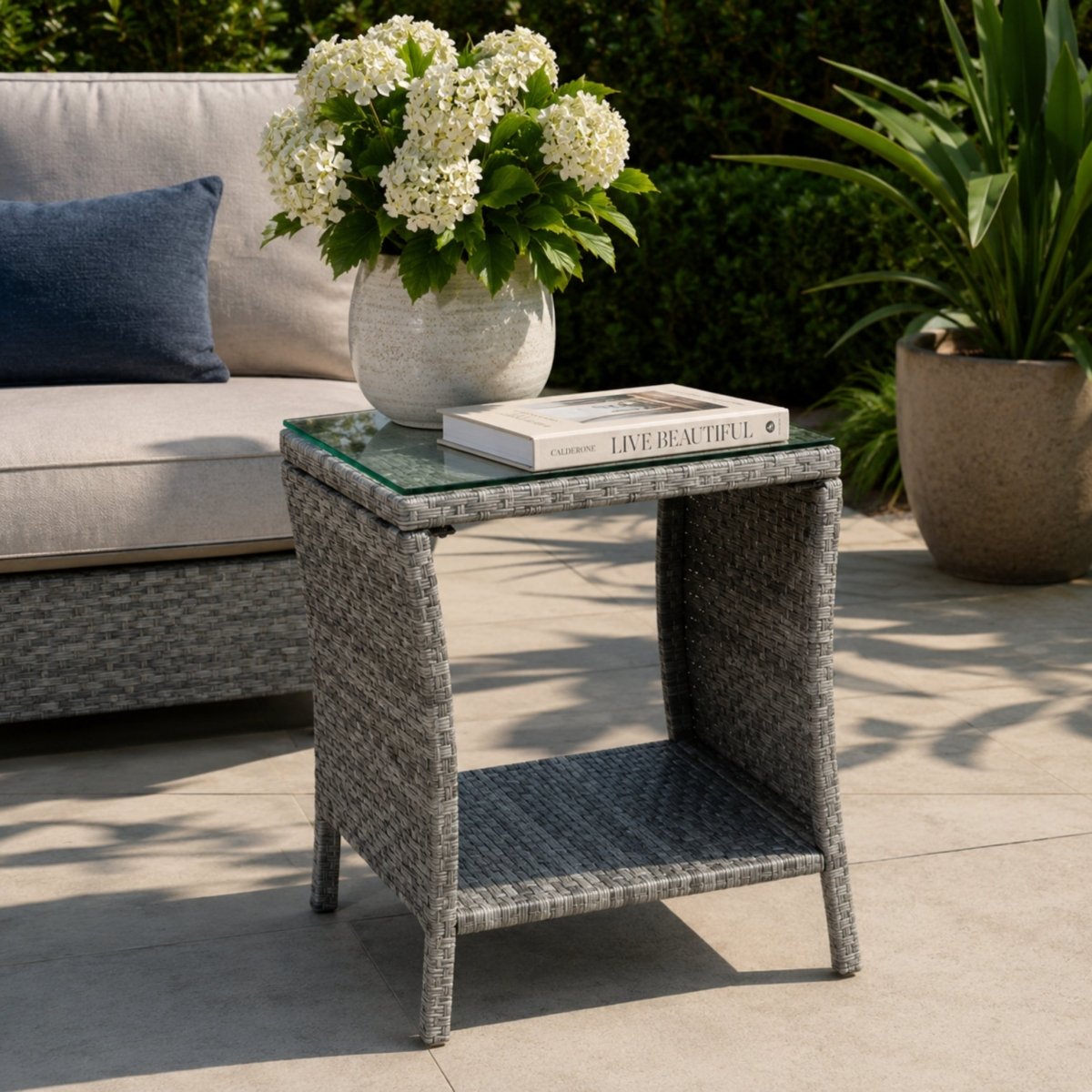 Detailed view of a grey woven all-weather outdoor table with a tempered glass top, decorated with a white floral vase and a coffee table book.