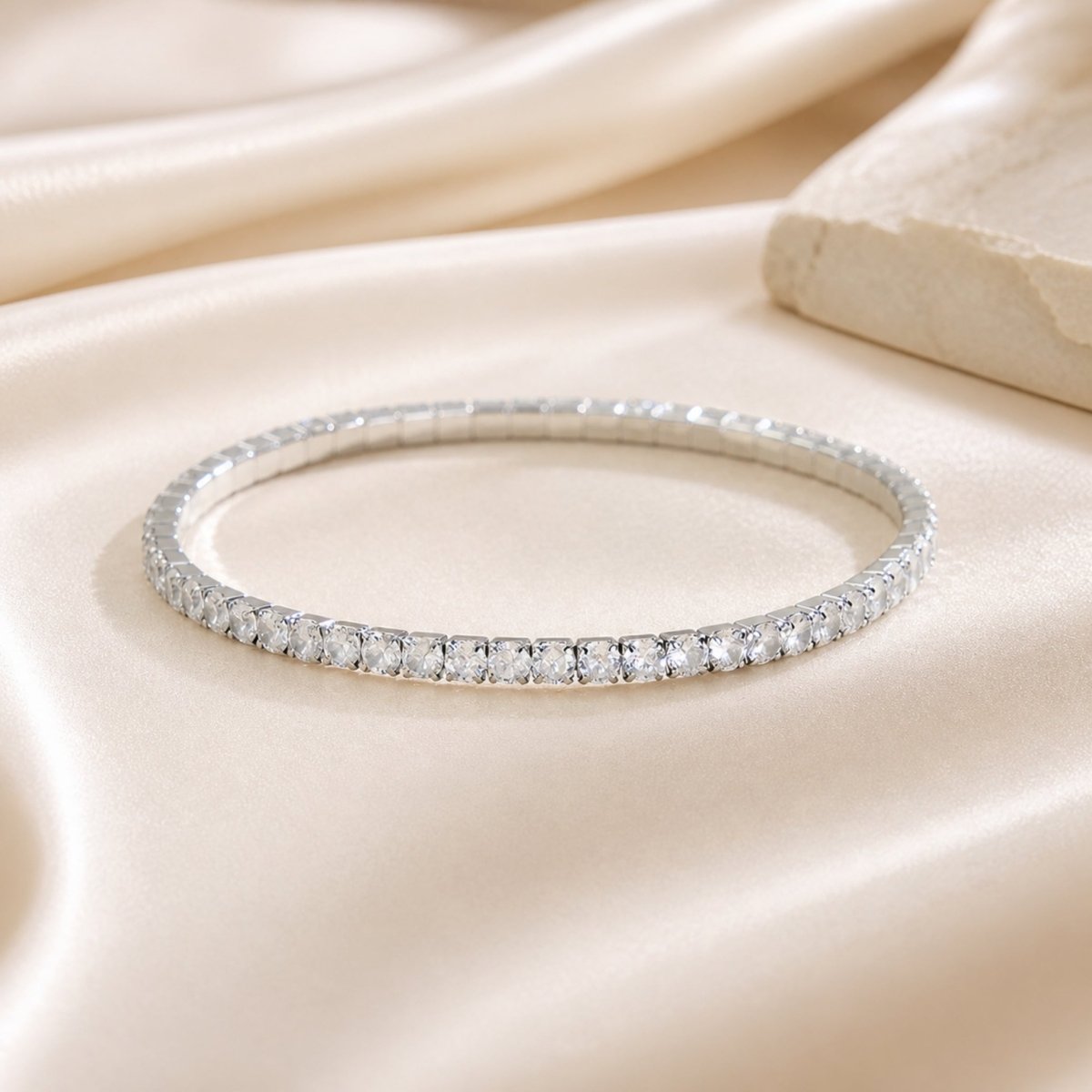 Close up macro shot of a dainty silver plated elastic tennis bracelet resting on a beige silk cloth next to a textured stone styling prop.