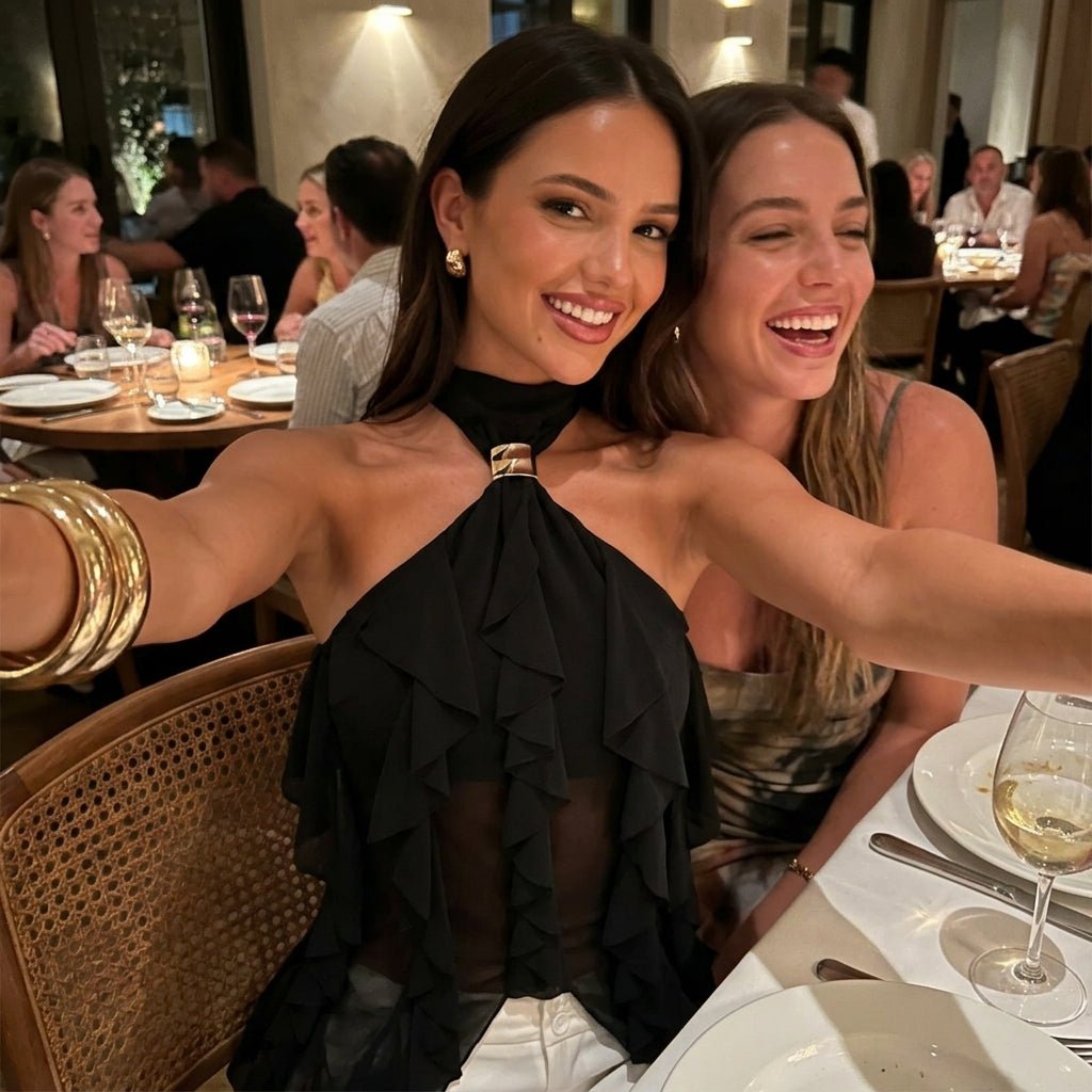 Two smiling women dining at a restaurant, with one taking a selfie wearing the Kouvr elegant black sheer ruffle halter evening top.