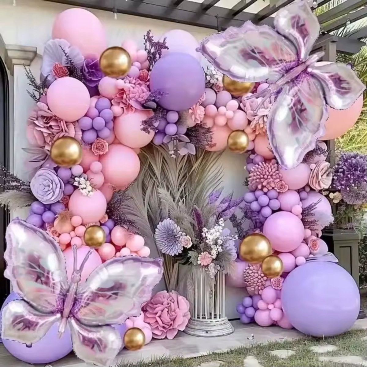 A sophisticated entryway balloon garland backdrop at a luxury property, combining matte lavender balloons, pink silk roses, gold chrome accents, and large 3D butterfly balloons over marble stairs.