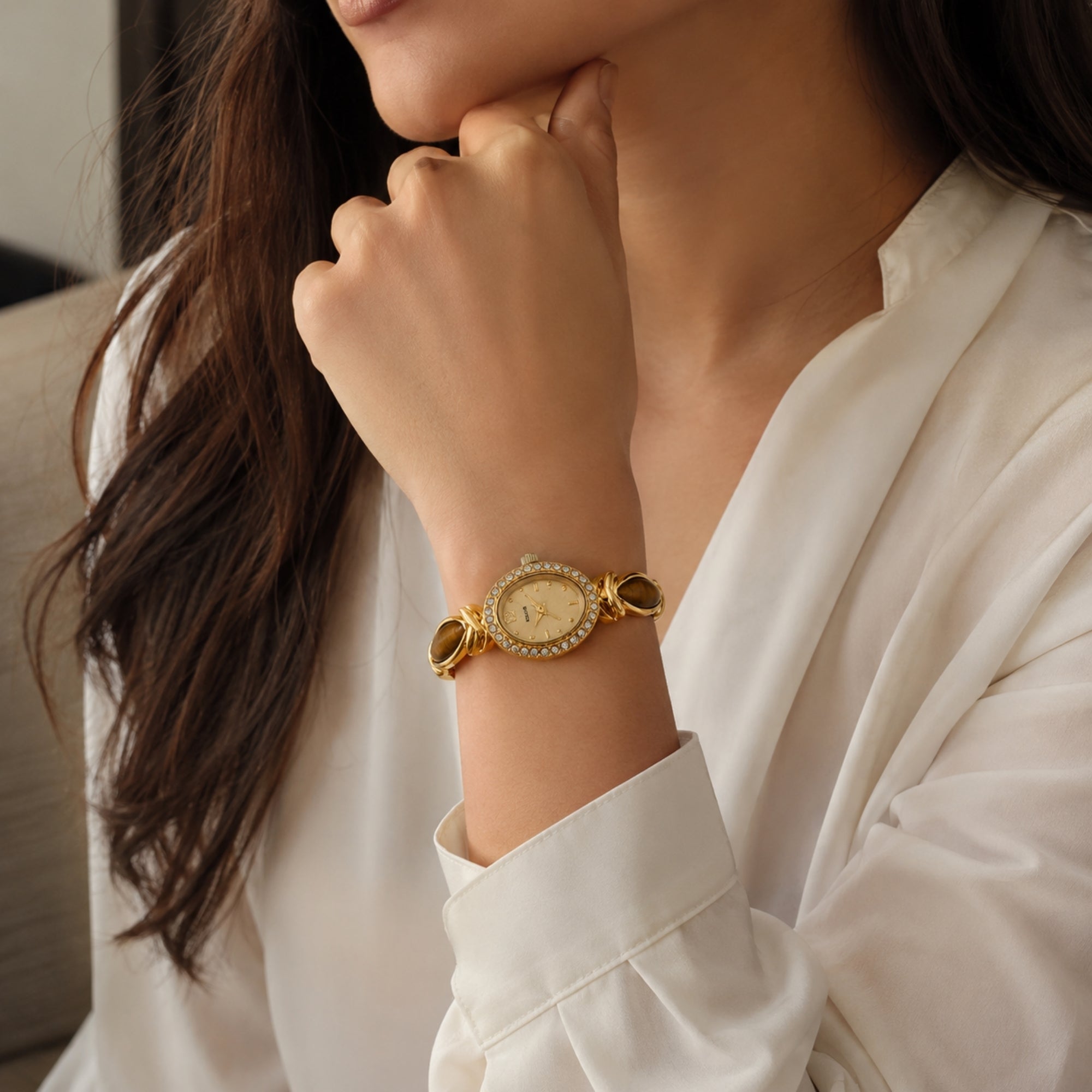 Close up of a woman resting her chin on her hand wearing a vintage gold and oval gemstone jewelry watch styled with a white silk blouse.