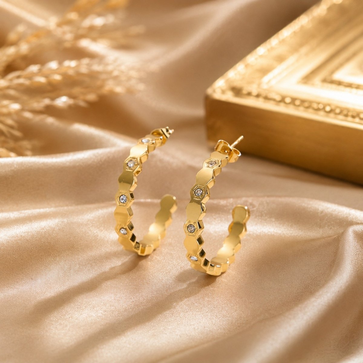 Fine gold geometric linked hoop earrings resting on luxurious draped champagne silk fabric next to an ornate gold vintage frame