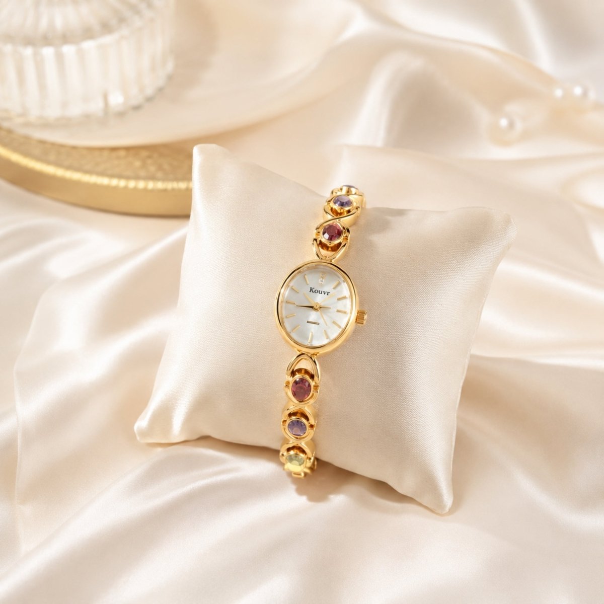 Close up product shot of the Kouvr gold ladies jewelry watch with red, purple, and green gemstones resting elegantly on a cream silk display cushion.