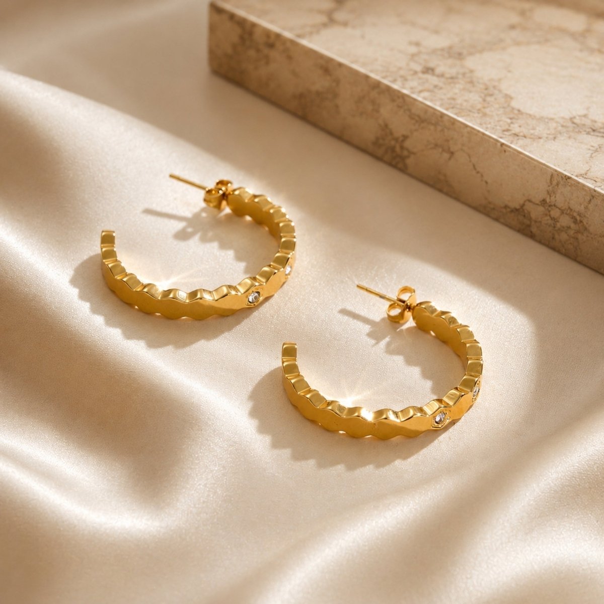 18k gold hexagon hoop earrings laid flat on soft cream silk fabric beside a textured travertine marble stone block