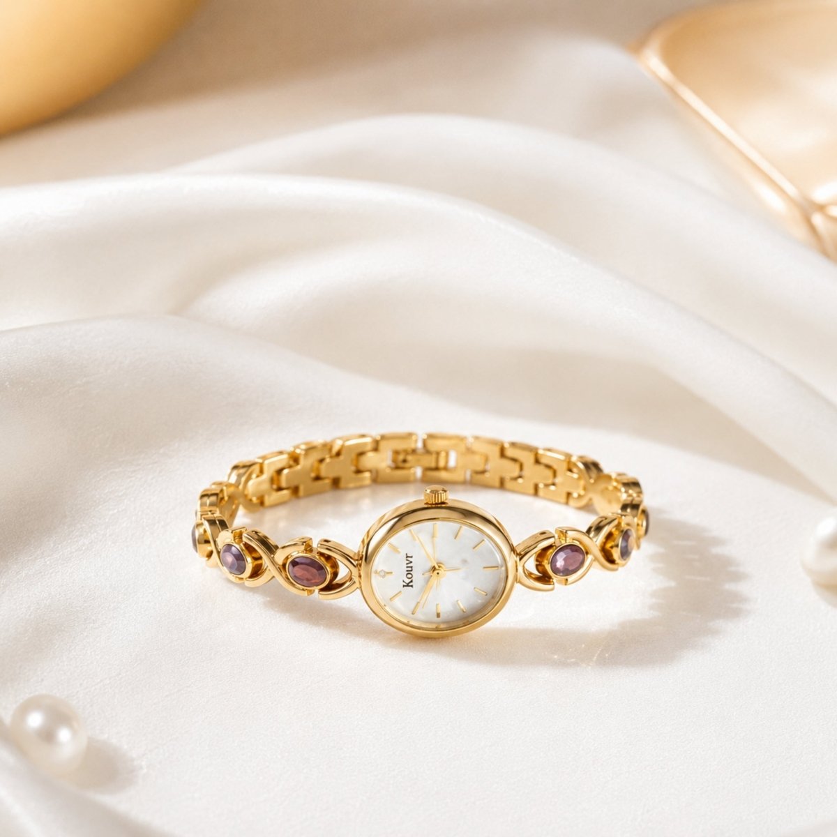 Horizontal flat lay of the gold Kouvr gemstone women's watch resting on draped white silk fabric, highlighting the elegant bracelet clasp and links.