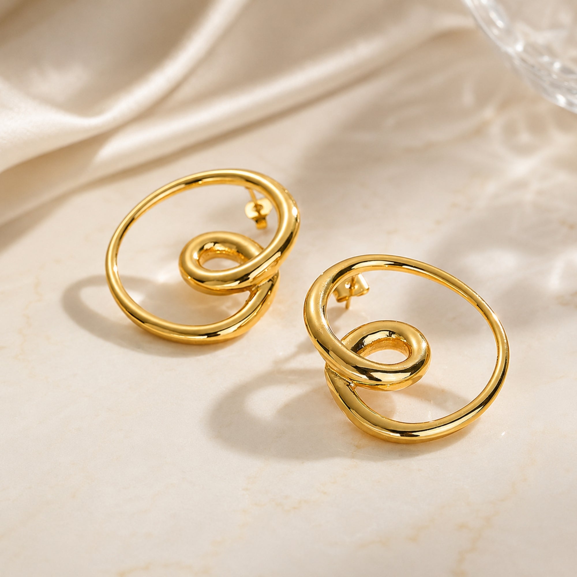 Two gold geometric knot stud earrings resting on a textured stone surface illuminated by warm sunlight and soft aesthetic shadows.