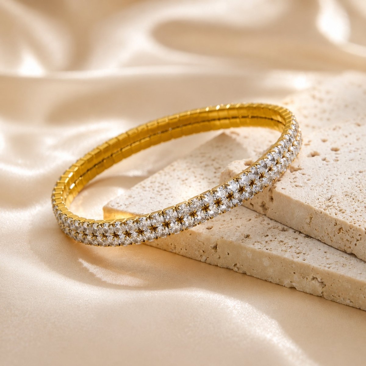 High end aesthetic flat lay featuring the Kouvr double row gold tennis bracelet resting elegantly on textured travertine stone blocks and smooth champagne satin fabric.