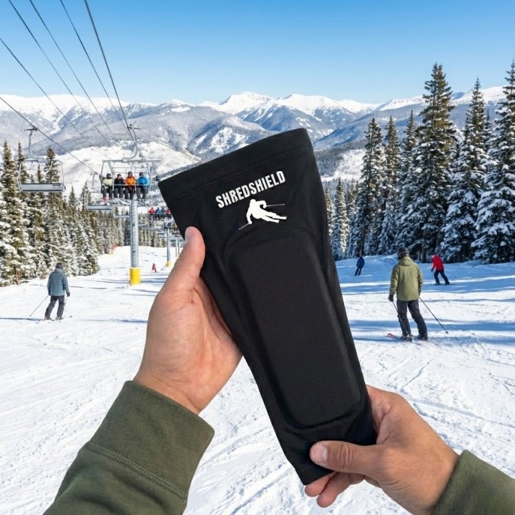 A first-person view of a hand holding up a black ShredShield ski shin pad, with a sunny ski resort, chairlift, and evergreen trees in the background.
