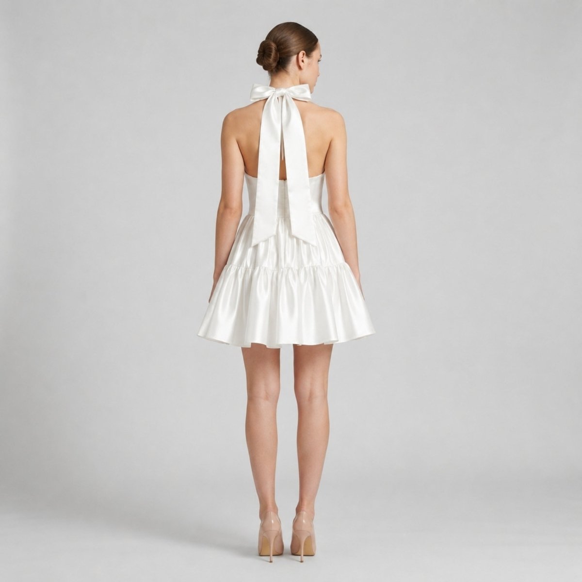Back view of a white satin open back dress featuring a large tie bow at the neck and a short ruffled skirt.