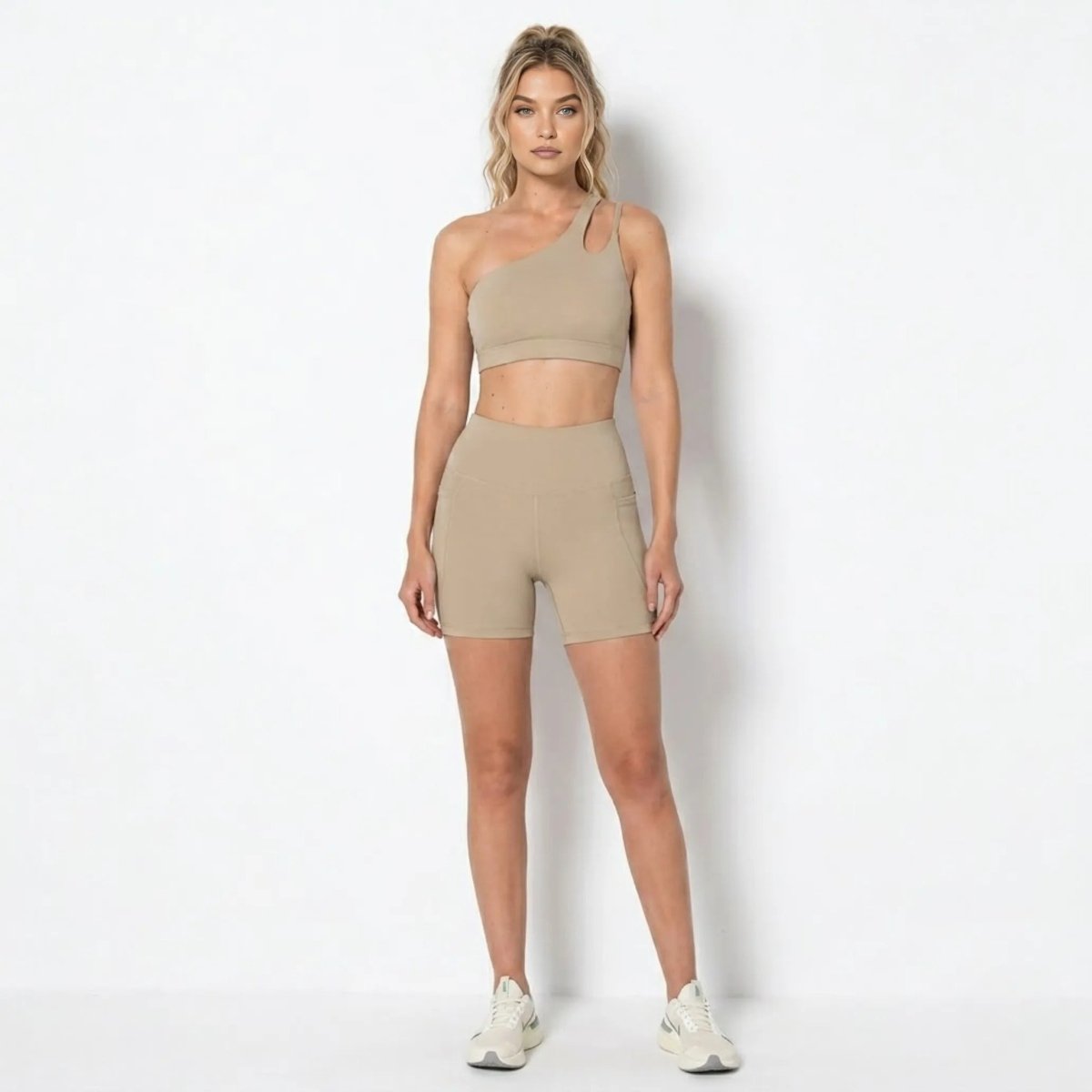 Blonde female model wearing Kouvr khaki asymmetrical one shoulder sports bra and matching high waisted biker shorts with pockets facing forward.