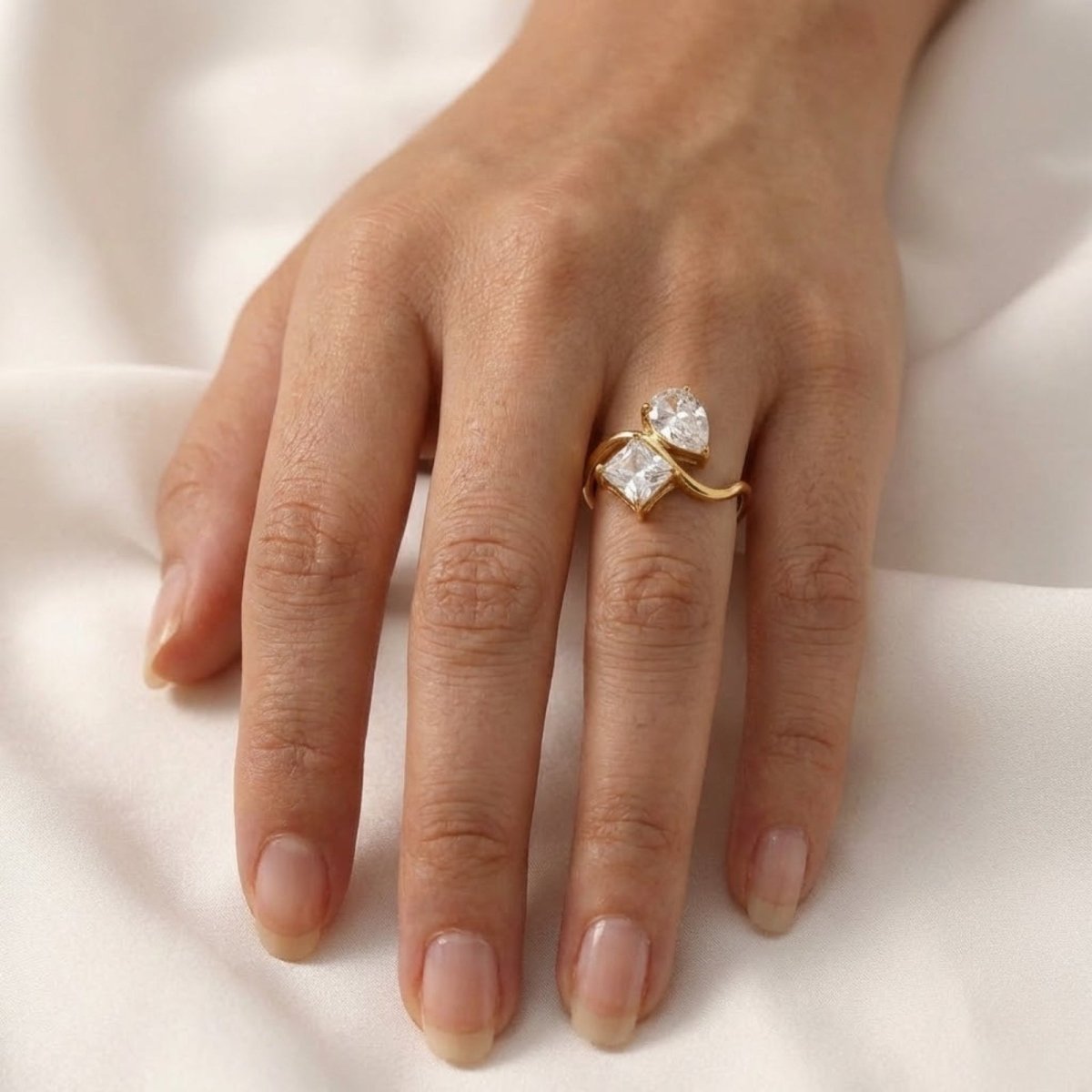 A top-down view of the Kouvr Blanca pear and princess cut ring on a woman's finger against a white fabric backdrop. 