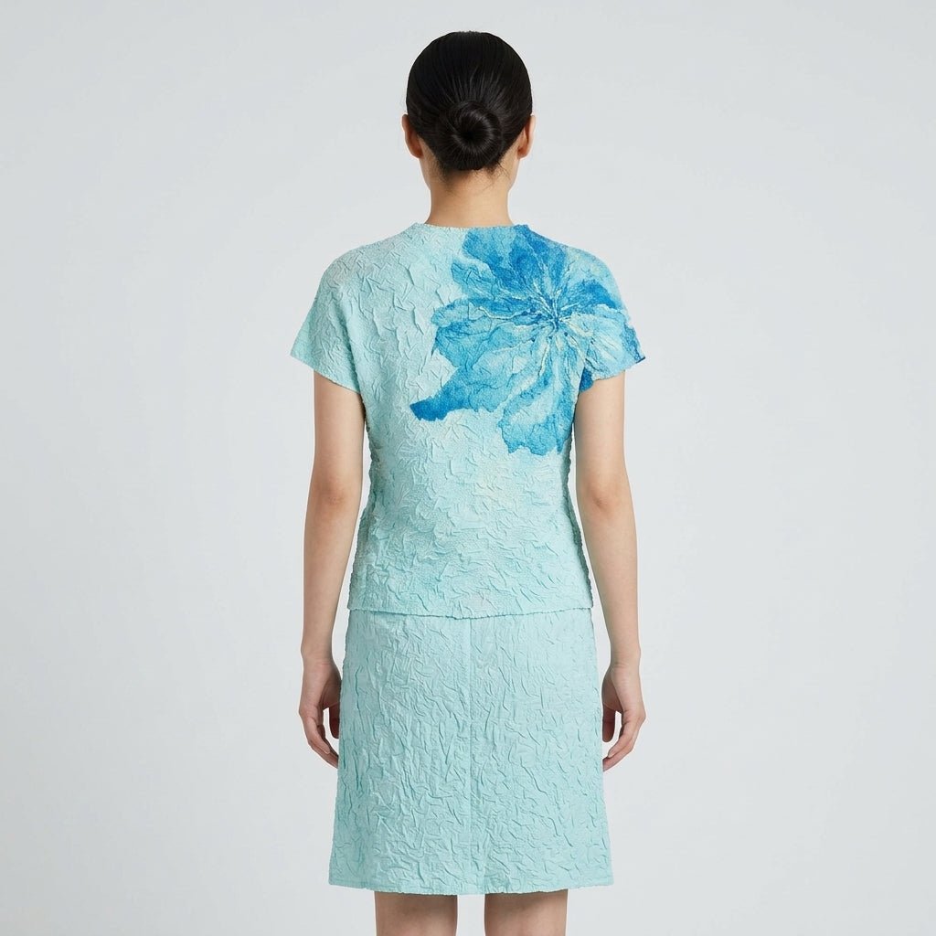 Back view of women's short sleeve light blue textured top showing the artistic wrapping floral shoulder print and matching skirt