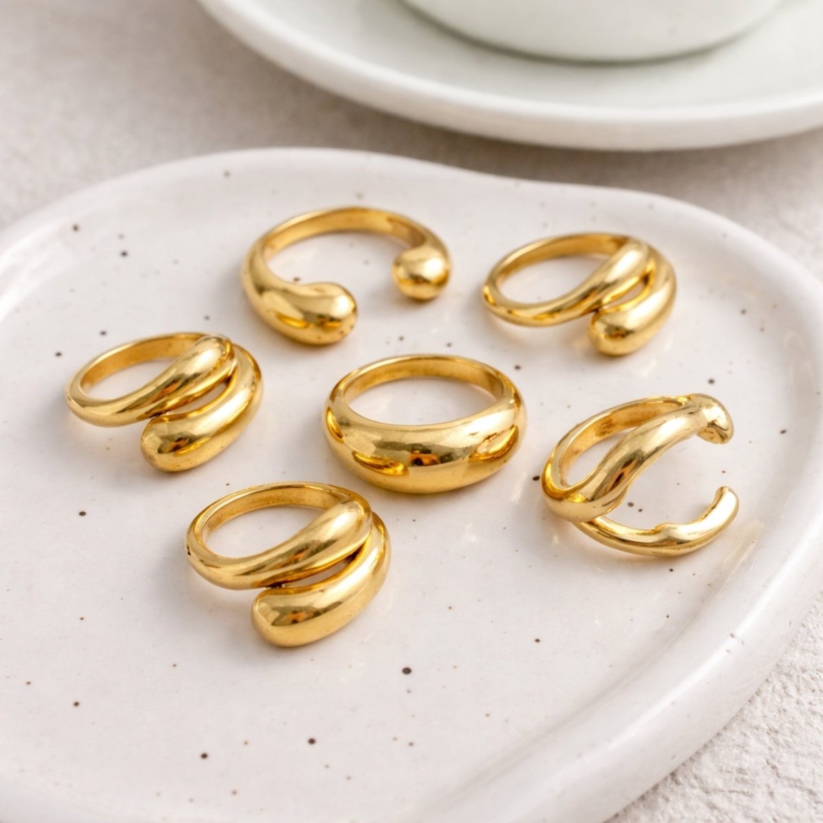 The six-piece Kouvr bold gold ring set displayed on a white ceramic tray, showcasing the variety of shapes including dome, crossover, and open-cuff rings.
