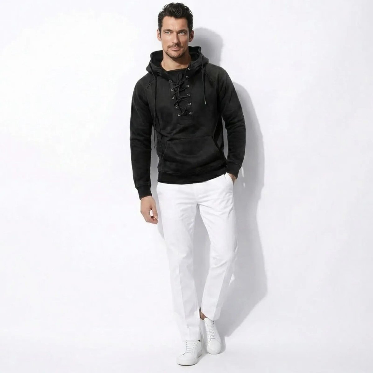 Male model wearing the Kouvr Cannon  washed black lace-up hoodie with white trousers and sneakers, standing against a white background.
