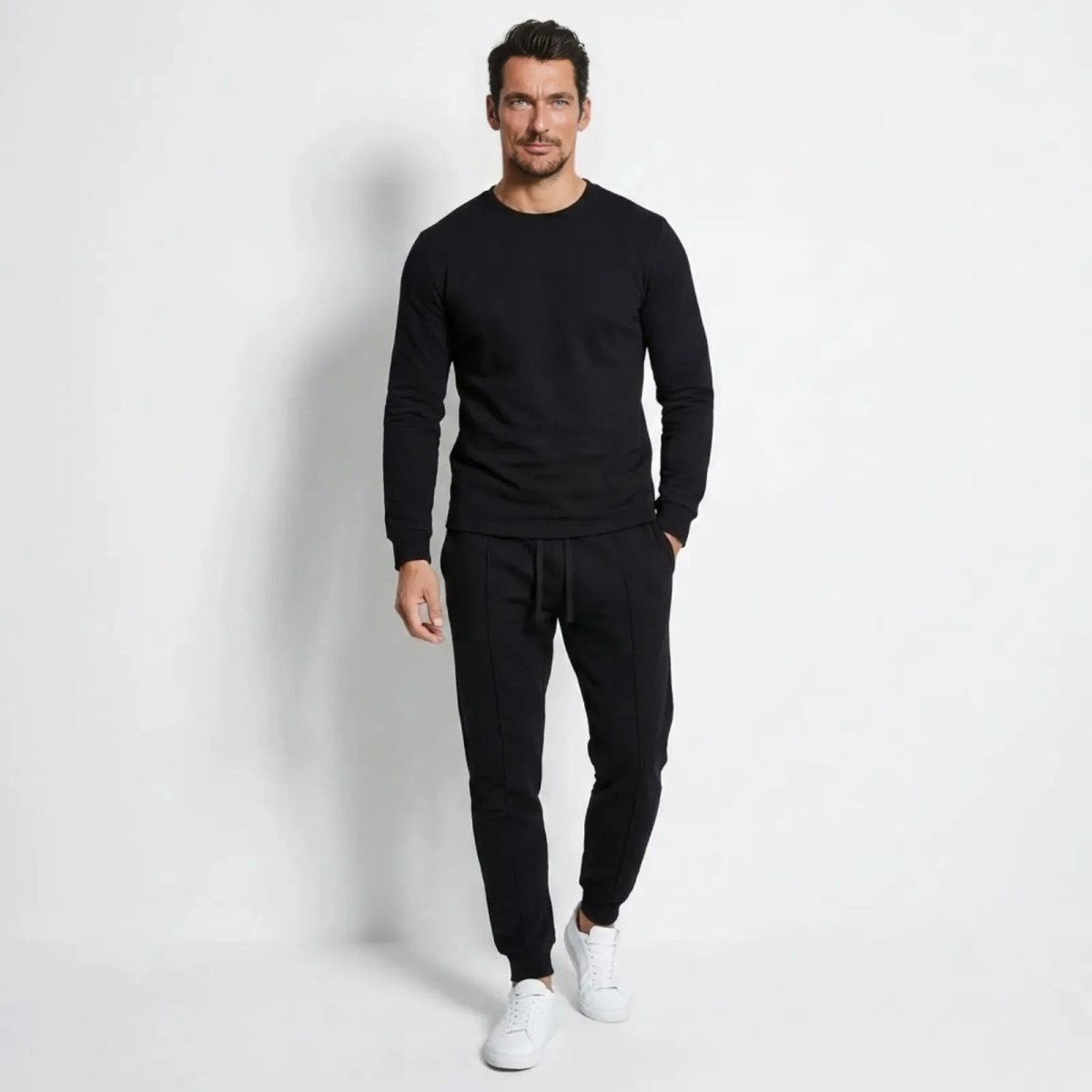 Man wearing the Kouvr Crue slim-fit black tracksuit set with a long-sleeve crewneck top and tapered joggers, styled with white sneakers.
