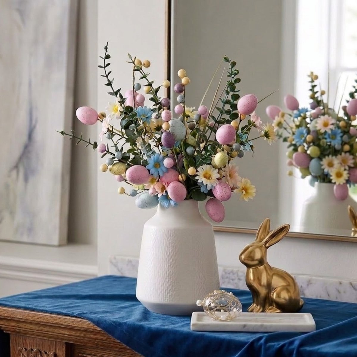 A tall white vase filled with a pastel Easter egg and daisy floral arrangement, sitting on a blue velvet runner next to a gold bunny statue.