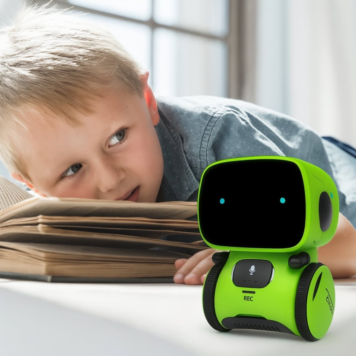 A young boy lies on the floor reading a book, looking with curiosity at the green Kouvr Fashion KAI interactive robot companion on the table next to him.