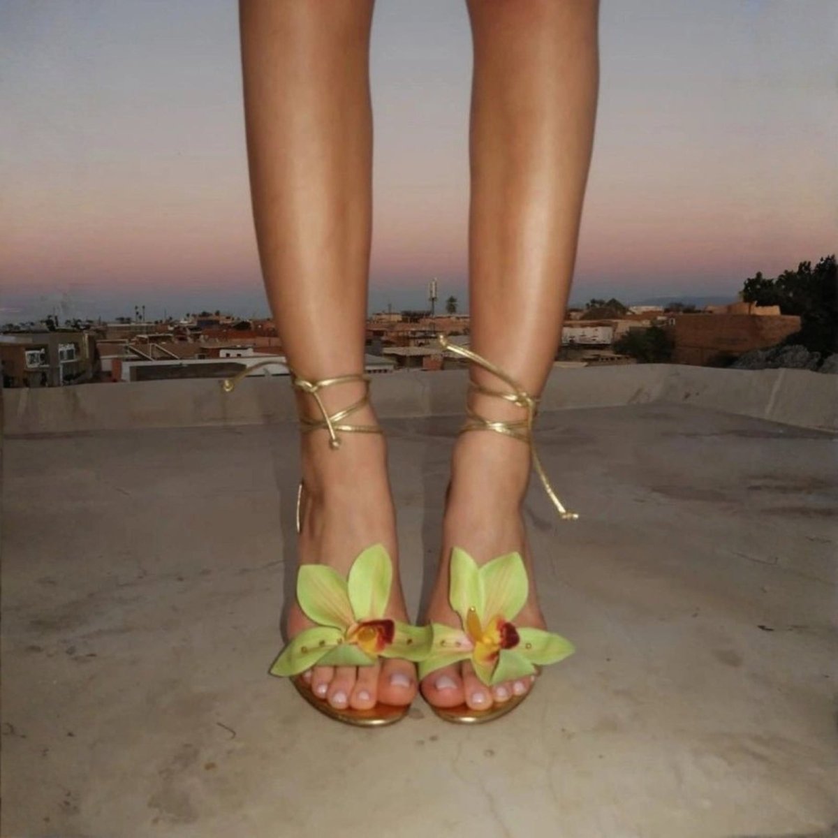 Woman wearing a high-heeled sandals with floral details on a rooftop at sunset.