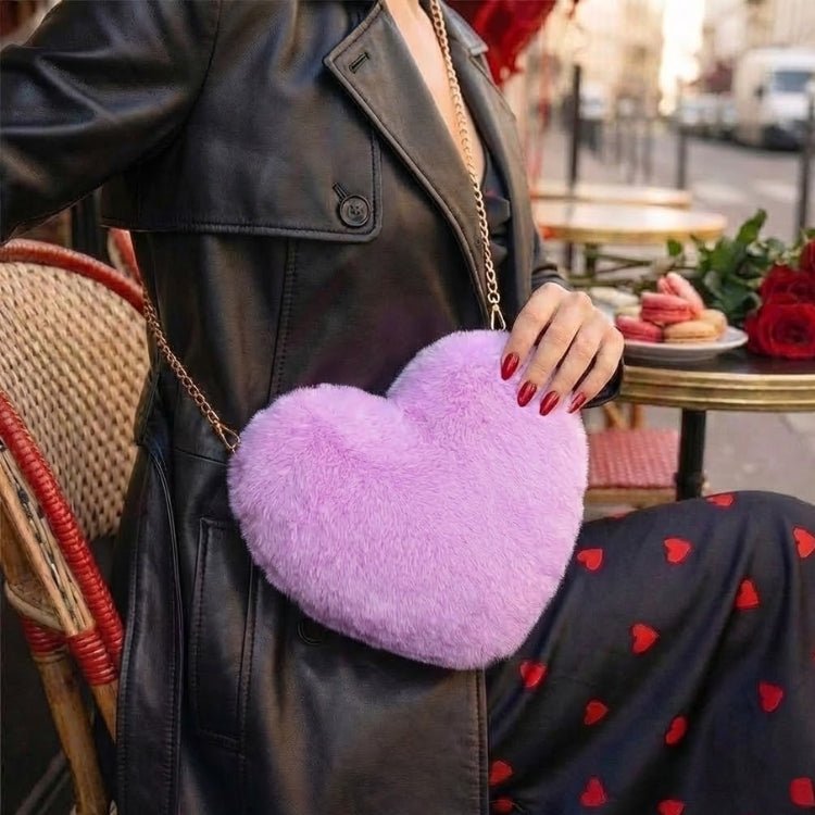 A stylish woman in a black leather coat sitting at a Parisian cafe table, wearing a lavender plush faux fur heart-shaped bag with a gold chain strap.

