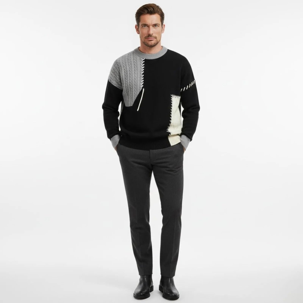 Male model facing forward wearing the Kouvr Fashion patchwork sweater in black, grey, and cream with dark grey trousers and black boots.
