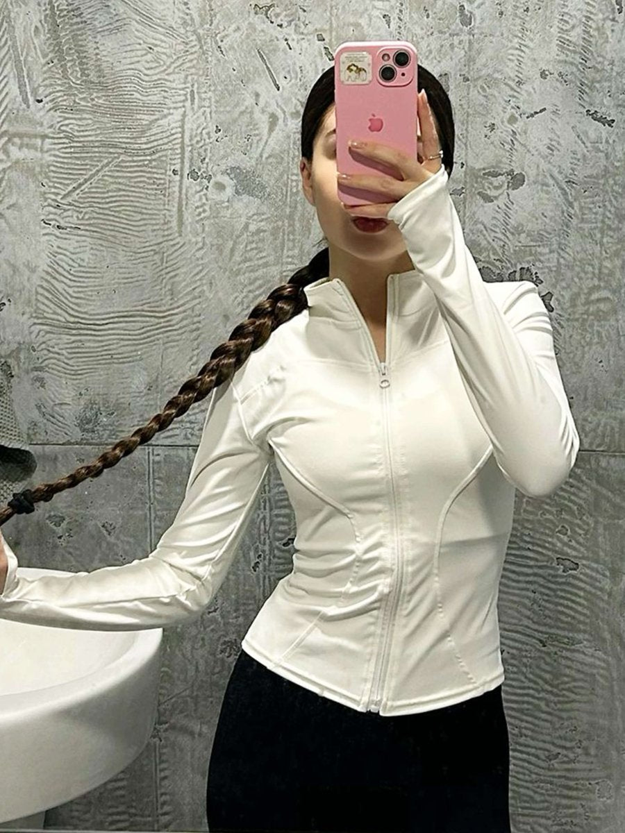 Woman with long blonde hair taking a mirror selfie in a gym wearing the white Nola Sculpt Athletic Jacket from Kouvr Fashion, showcasing its form-fitting design.