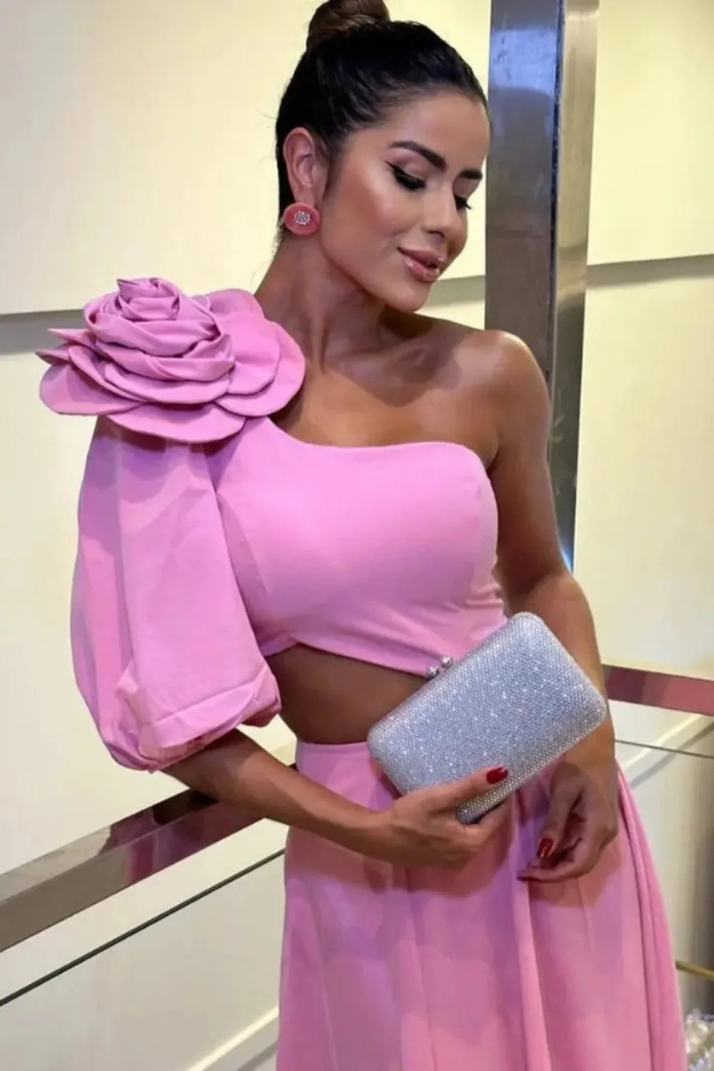 Model wearing Kouvr's pink one-shoulder midi dress with a large rose on the shoulder and a side waist cutout, standing by a pool at night