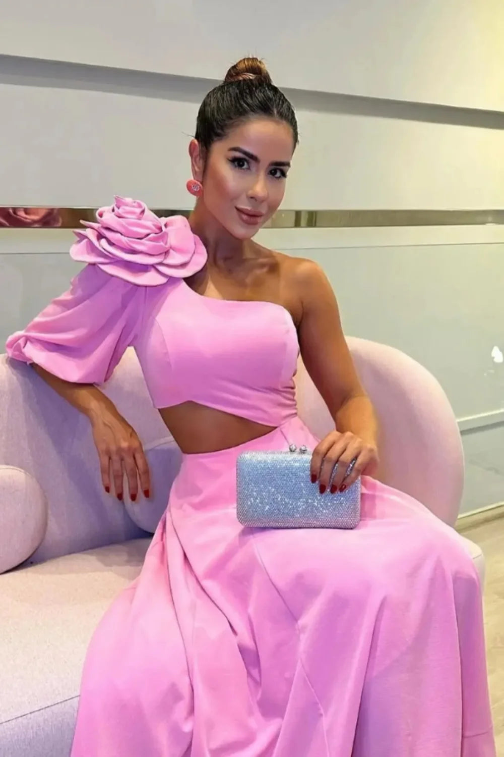 Model wearing Kouvr's pink one-shoulder midi dress with a large rose on the shoulder and a side waist cutout, standing by a pool at night