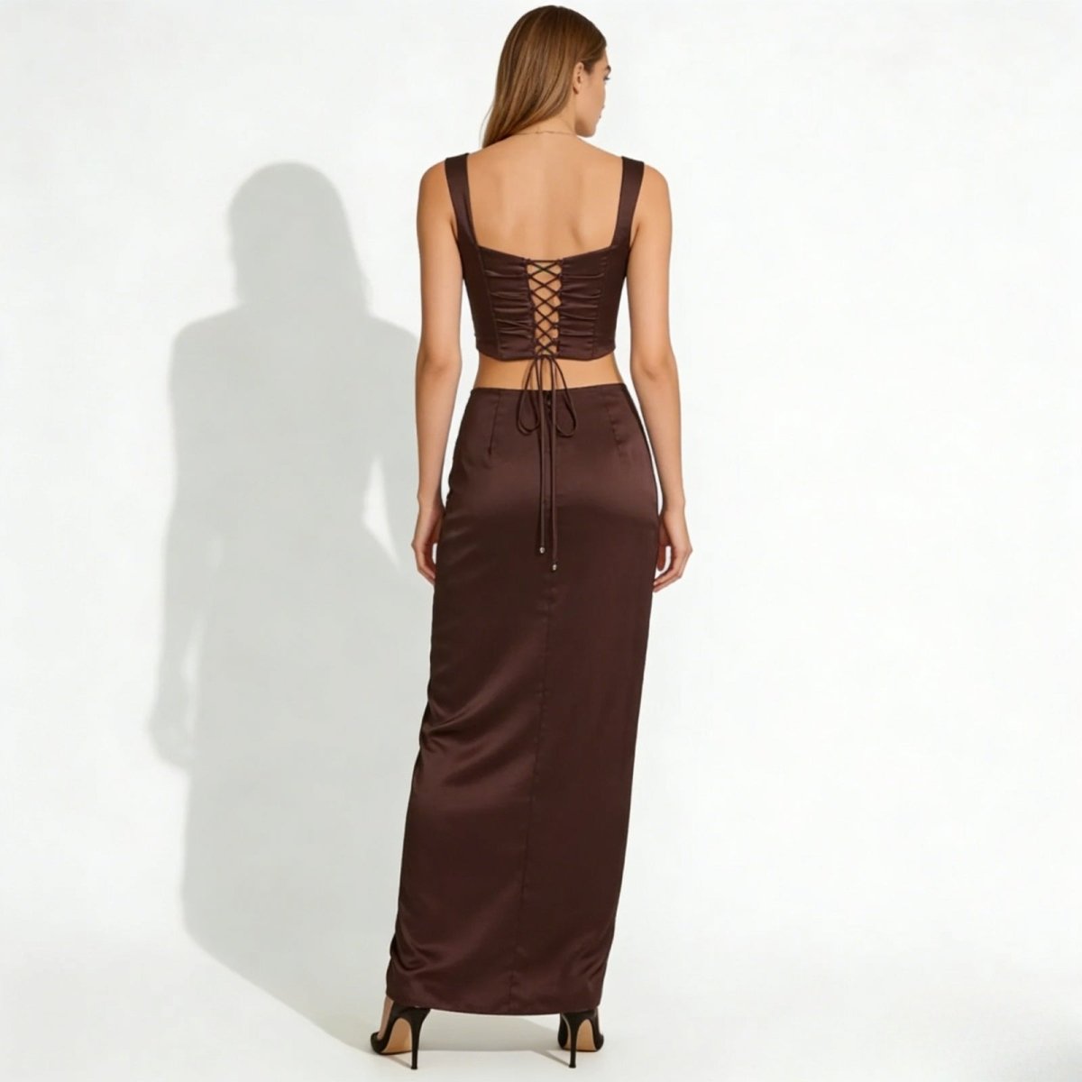 Back view of a model in the rona dark brown satin set, detailing the adjustable lace-up corset back of the top and the full length of the maxi skirt.