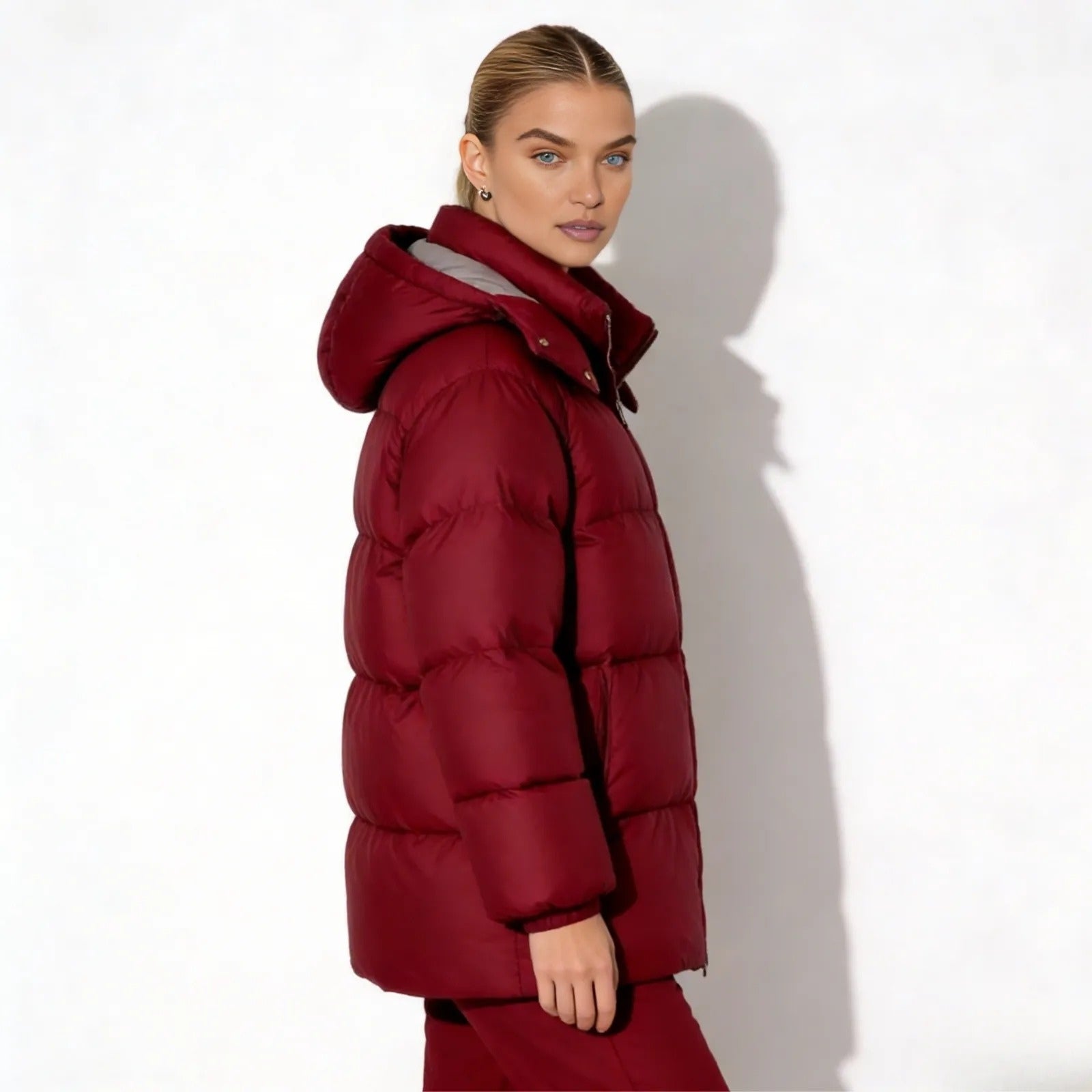 Side profile of a woman wearing the Kouvr Fashion mid-length hooded puffer coat in burgundy showcasing its oversized silhouette. 