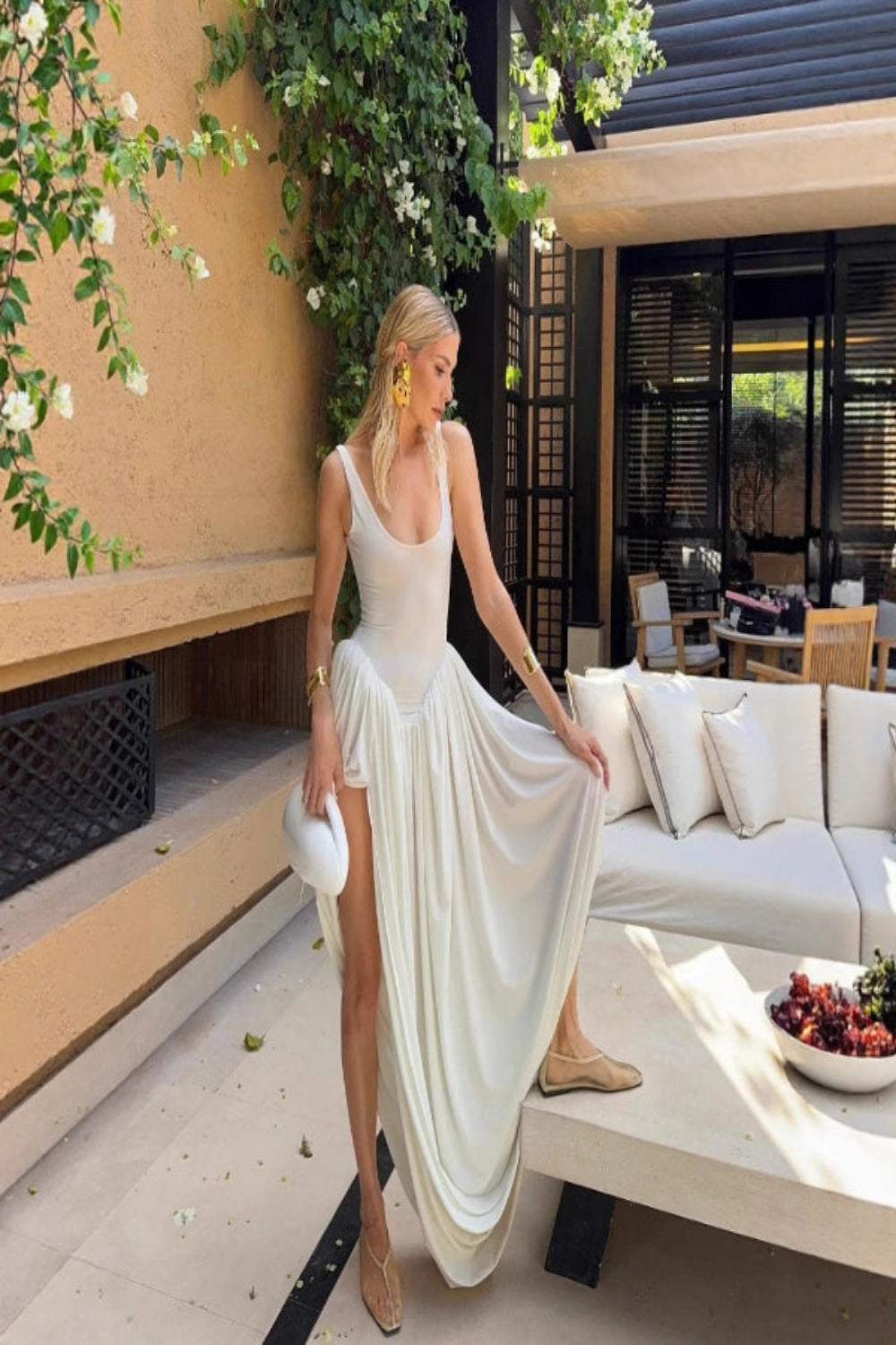 Woman with a brown bob hairstyle wearing the Kouvr Stacey white draped maxi dress with gold cuffs and a yellow clutch bag