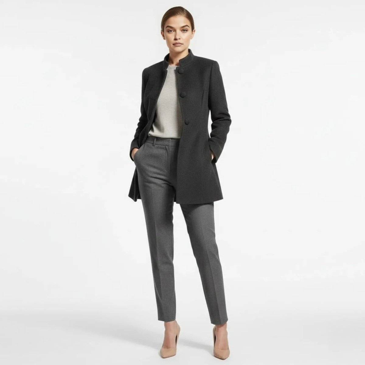 Model wearing the Kouvr dark grey funnel neck wool blend coat with her hands in the pockets, styled with matching grey trousers and nude heels for a professional look. 
